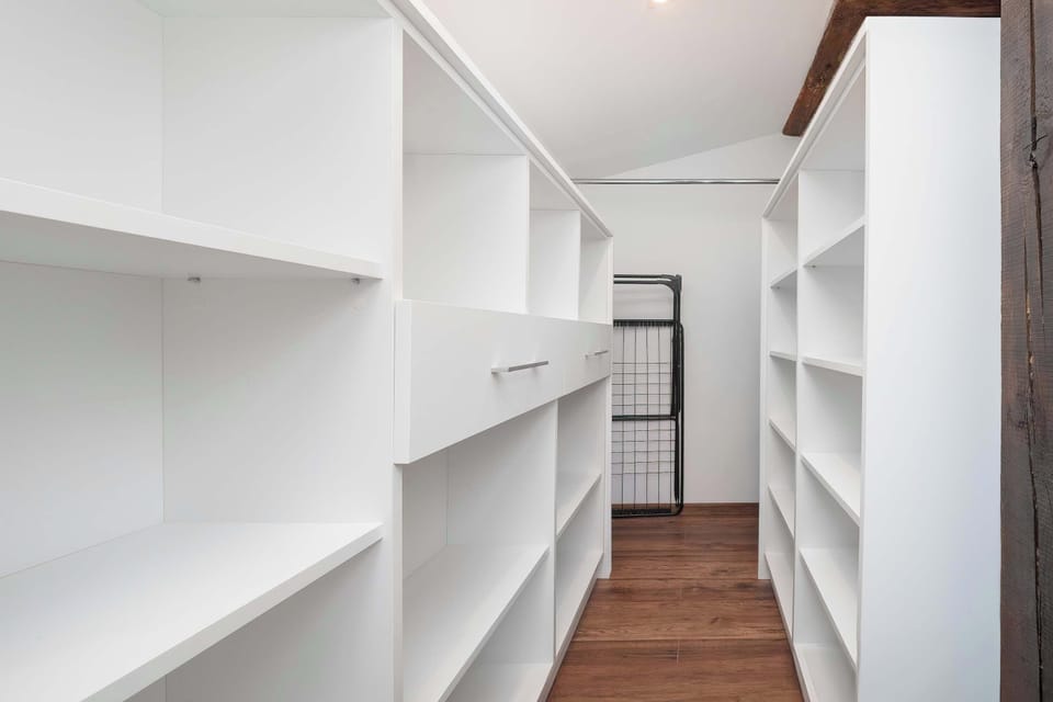 dedicated walk-in closet or storage room is lined with custom-built white shelving units on both sides. The narrow space features warm wood flooring and leads to a clothes rack at the far end.
