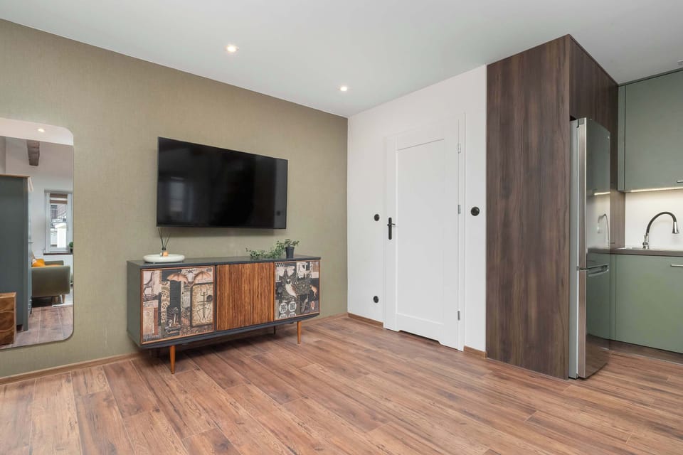A wall-mounted television hangs above a vintage-style wooden sideboard with colorful drawer fronts. The room features warm wood flooring and a doorway leading into the adjoining kitchen space.