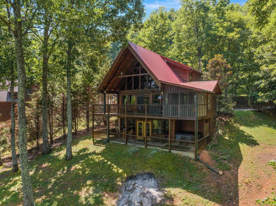 A charming A-frame cabin with a striking red metal roof nestled among towering trees, featuring wraparound decks and a private hot tub, offering a peaceful woodland retreat.