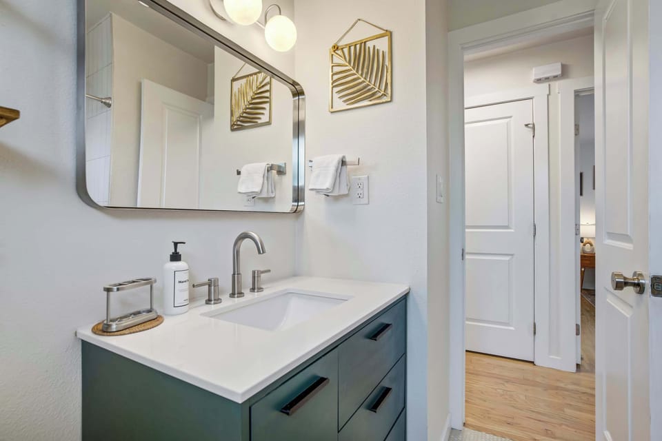 Green shaker cabinetry and a white quartz counter anchor the vanity, framed by a sleek mirror, brass sconce lighting, and open wood shelving that adds a touch of warmth to the crisp white walls.
