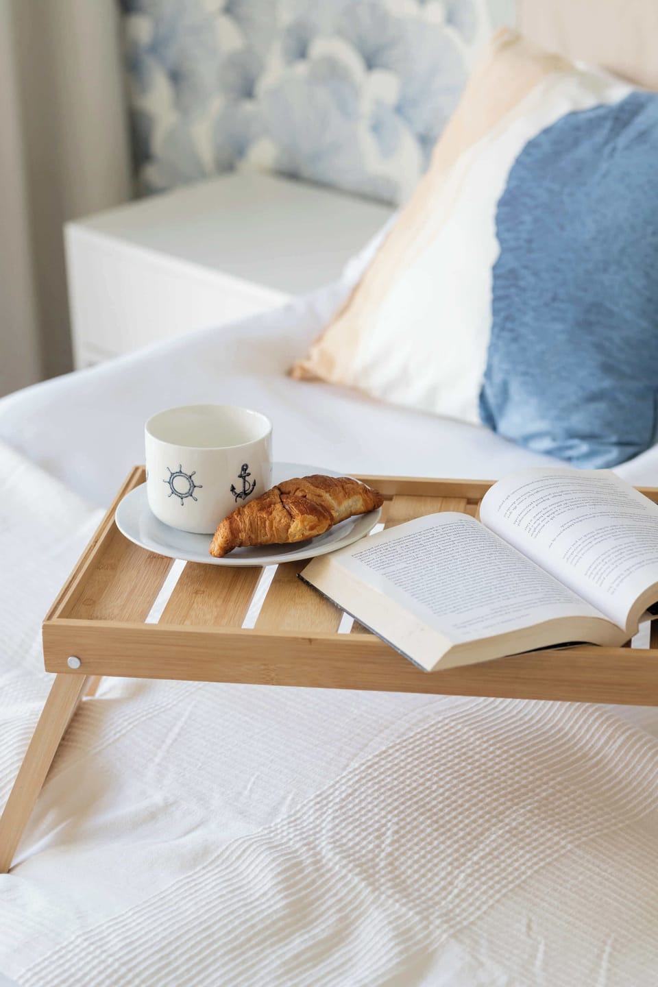 A close-up view focuses on the wooden breakfast tray laid out with a white coffee mug, a croissant, and an open book on the bed.