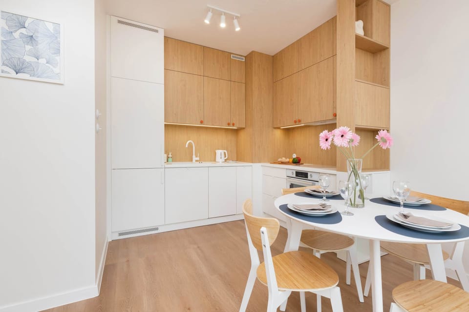 A round white dining table is set with plates and decorated with a vase of pink flowers. The wood-grained kitchen cabinets frame the background.