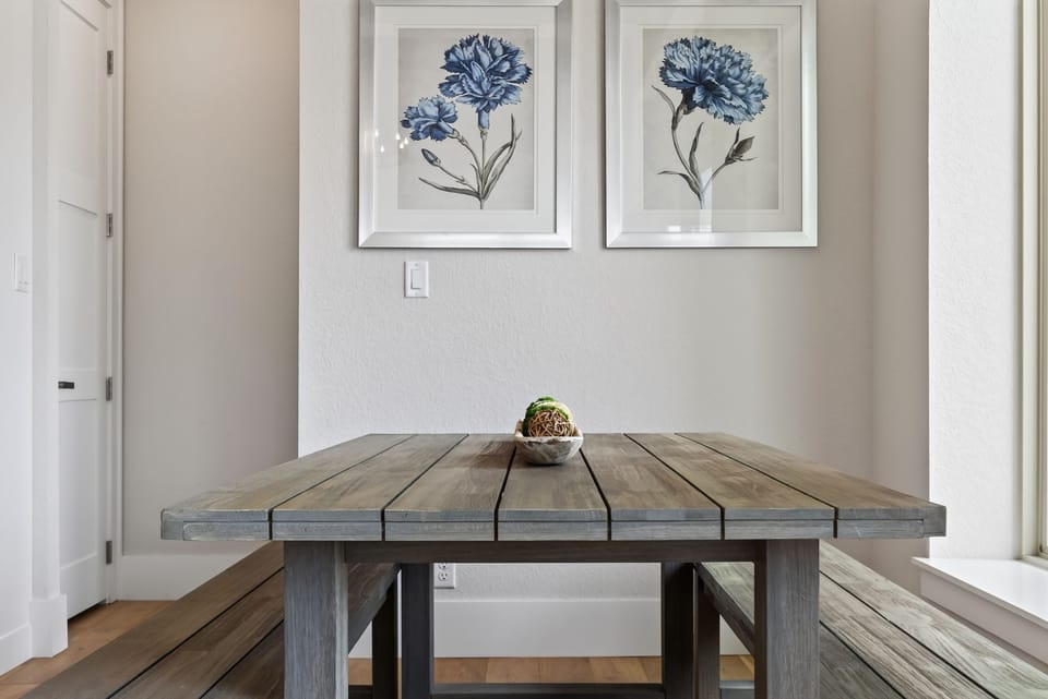 A slatted wooden dining table and matching bench set beneath framed blue floral prints.