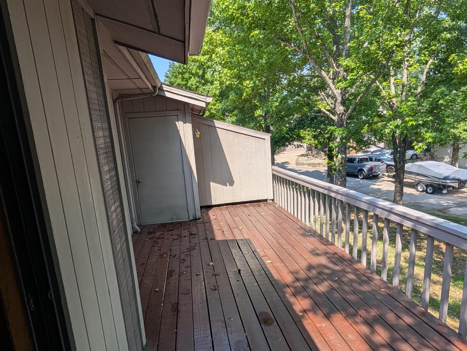 Wood-plank balcony with gray railing overlooks leafy trees, parked vehicles, and a covered boat.