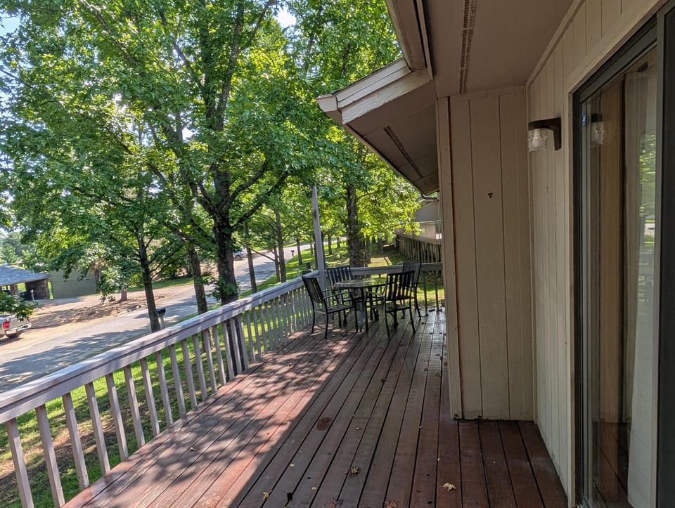 Spacious wood-plank balcony with gray railing and black metal table set, shaded by leafy green trees.
