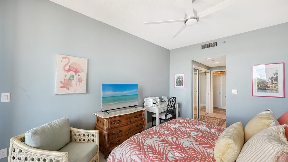 Bedroom with coral and gray leaf-patterned bedding, wicker armchair, wooden dresser with TV, and white desk.