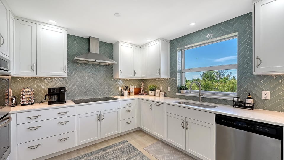 The kitchen features white cabinetry, a stainless steel range hood, green herringbone tile backsplash, and a window with a garden view.