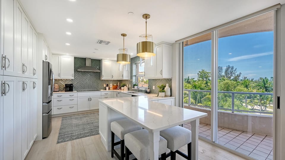 White quartz island with upholstered stools sits next to sliding glass doors opening to a balcony with tree views.