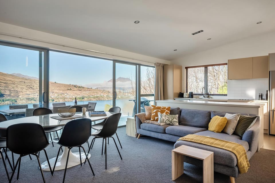 The warm, welcoming heart of the home: a plush sectional sofa, generous dining table, and a well-appointed kitchen that opens straight onto the BBQ deck and glorious lake view beyond.