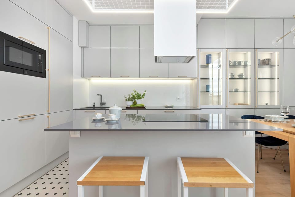 A bright kitchen island stands in the center of the space with two wooden stools tucked neatly underneath. The minimalist design highlights seamless white cabinetry, high-end integrated appliances, and a smooth grey countertop.