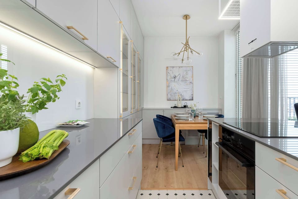 A narrow kitchen corridor looks ahead toward a small dining table paired with deep blue chairs. Sleek white lower cabinets with dark countertops line the left side under soft under-cabinet LED strip lighting.