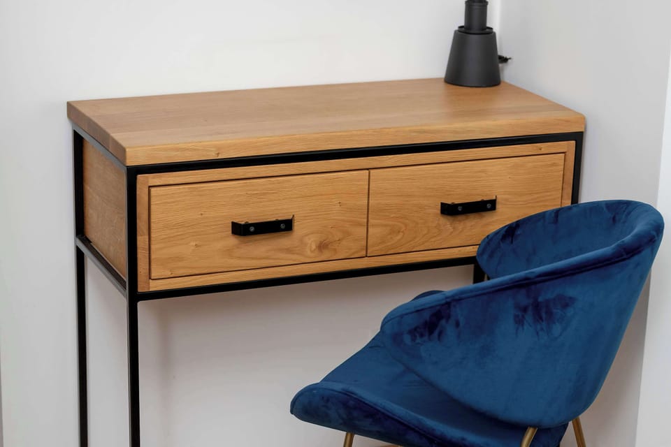 A compact wooden desk with two drawers is paired with a single deep blue velvet chair. A simple black lamp sits on top of the desk surface against a clean, unadorned white wall.