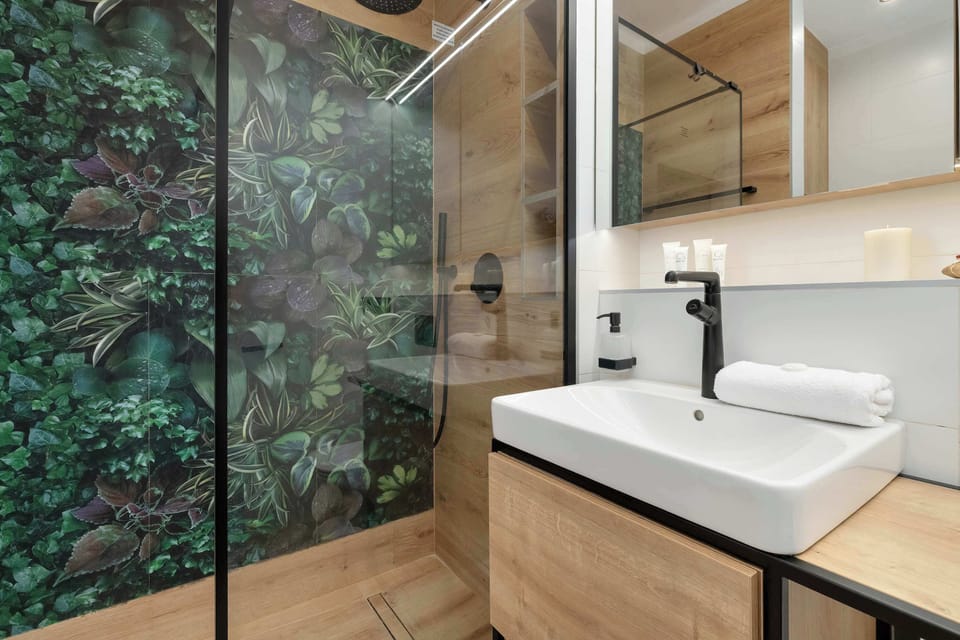 A modern bathroom vanity setup displays a rectangular white sink resting on a light wood counter with a black faucet. A vibrant green foliage wall panel is visible behind the glass shower screen, adding a natural touch.