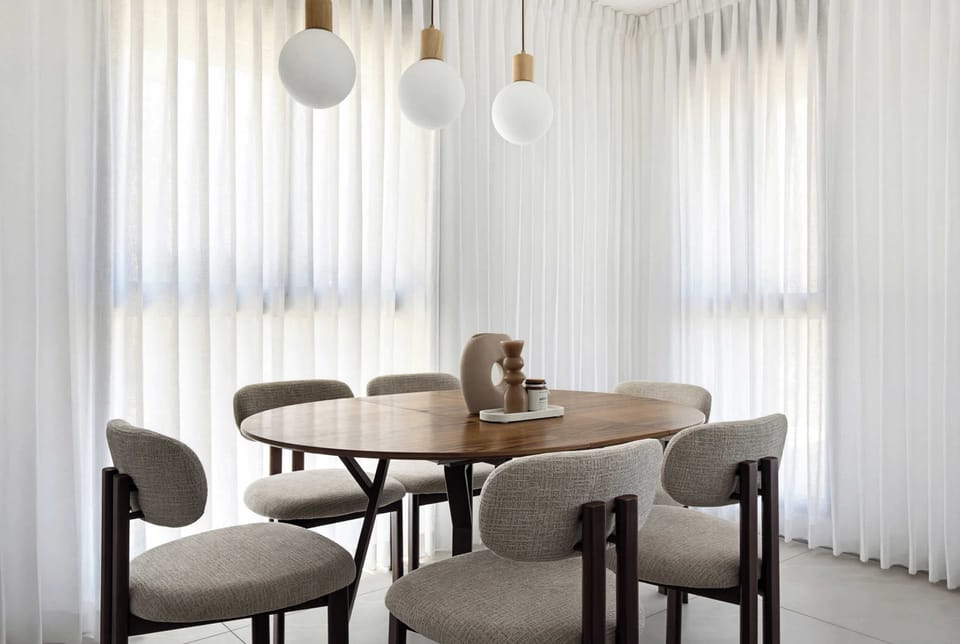 Elegant dining setting features sculptural pendant lights, modern chairs, natural wood table, and flowing lightweight curtains overhead.
