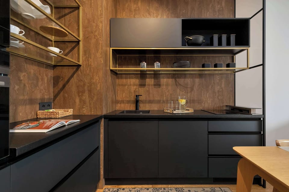 A sleek minimalist kitchen boasts matte black cabinetry paired with warm wood-grained walls. Open brass shelves hold glasses and kitchenware.