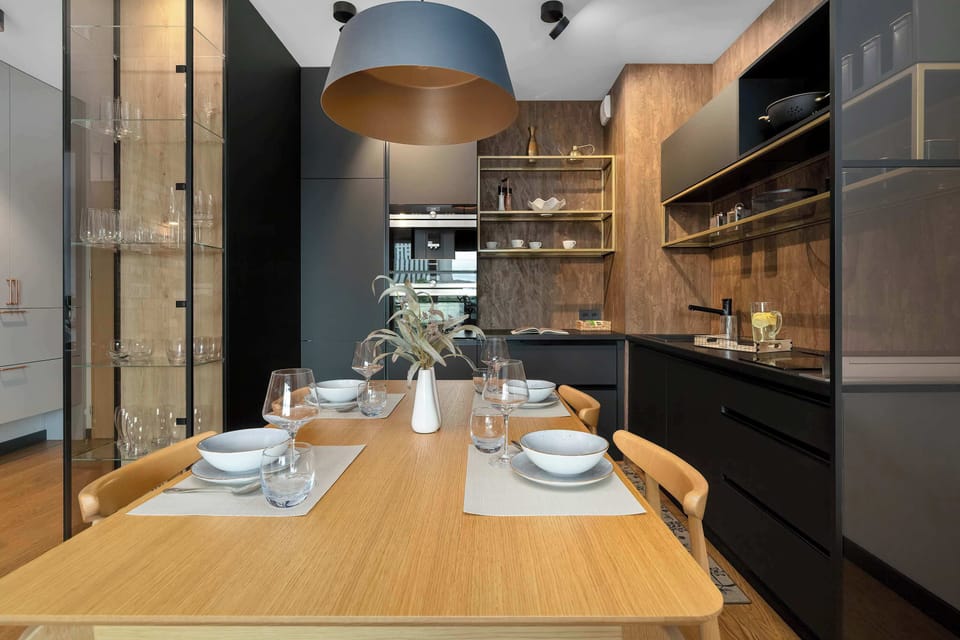 A straight-on perspective displays the rectangular wooden dining table prepared for four guests. The dark kitchen and a glass display cabinet frame the sides.