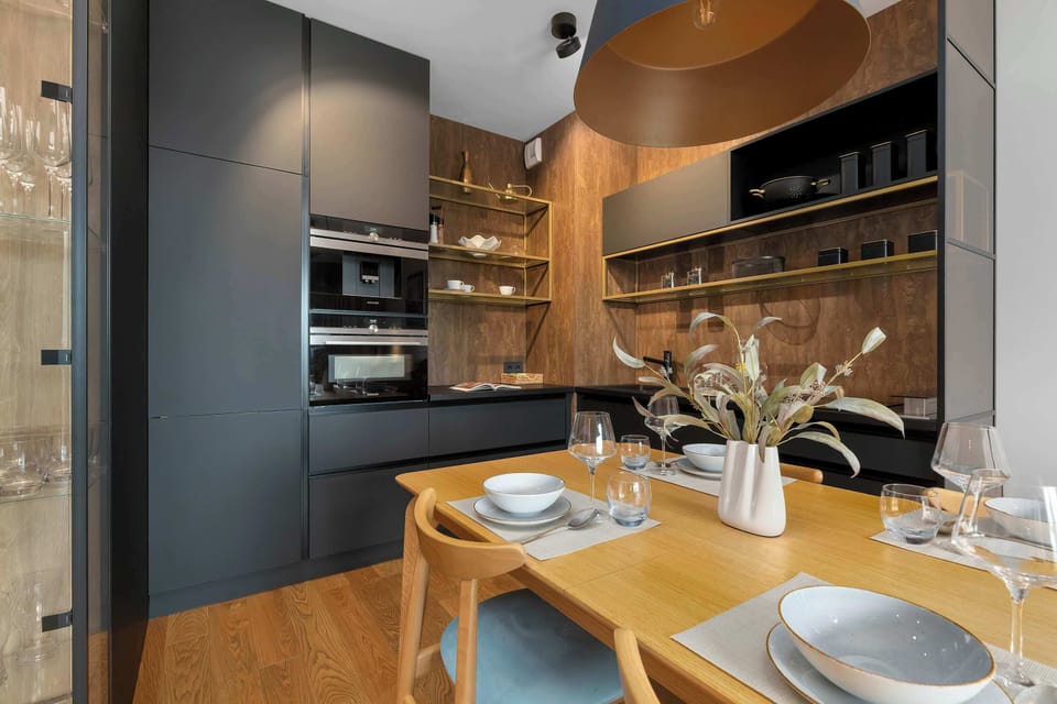 Looking across the dining table, the view reveals integrated kitchen appliances inside sleek black cabinetry. Warm wooden floors unify the space.