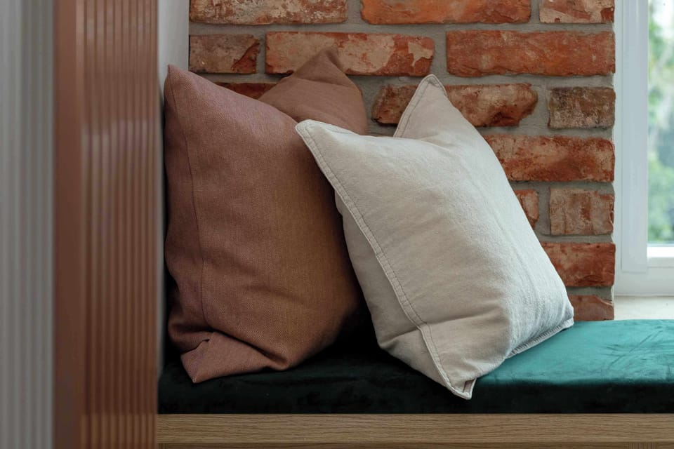 A close-up focuses on a pair of decorative pillows - one rust-colored and one off-white - resting on a green velvet window bench against a brick wall.