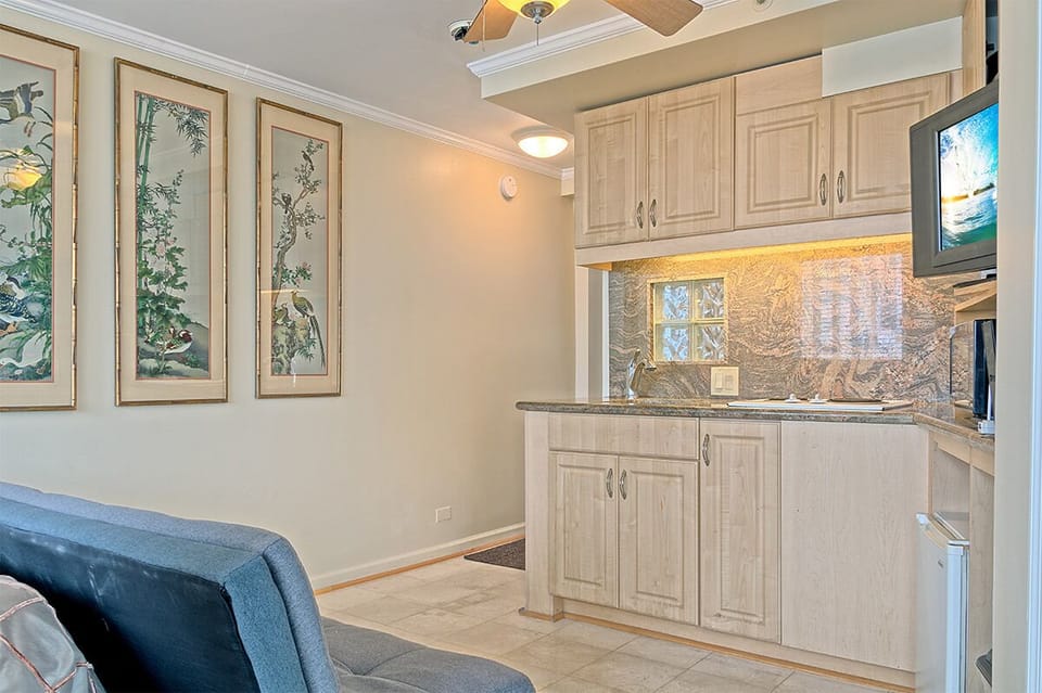 Your open-concept living space blends tropical artistry with modern comfort—ceiling fan above, full kitchenette ready for meals, and soft lighting welcoming you home.