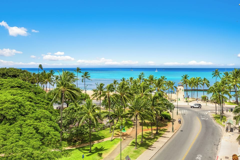 A scenic coastal property location features palm-lined streets leading to a pristine beachfront with turquoise waters and distant sailboats under clear blue skies.