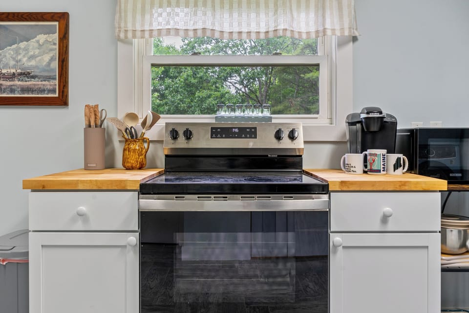 This close-up view details the centered GE electric range and its smooth glass cooktop. Thick wood butcher block countertops flank the oven, holding provided cooking utensils, a complete knife block, and a Keurig coffee station with curated mugs.
