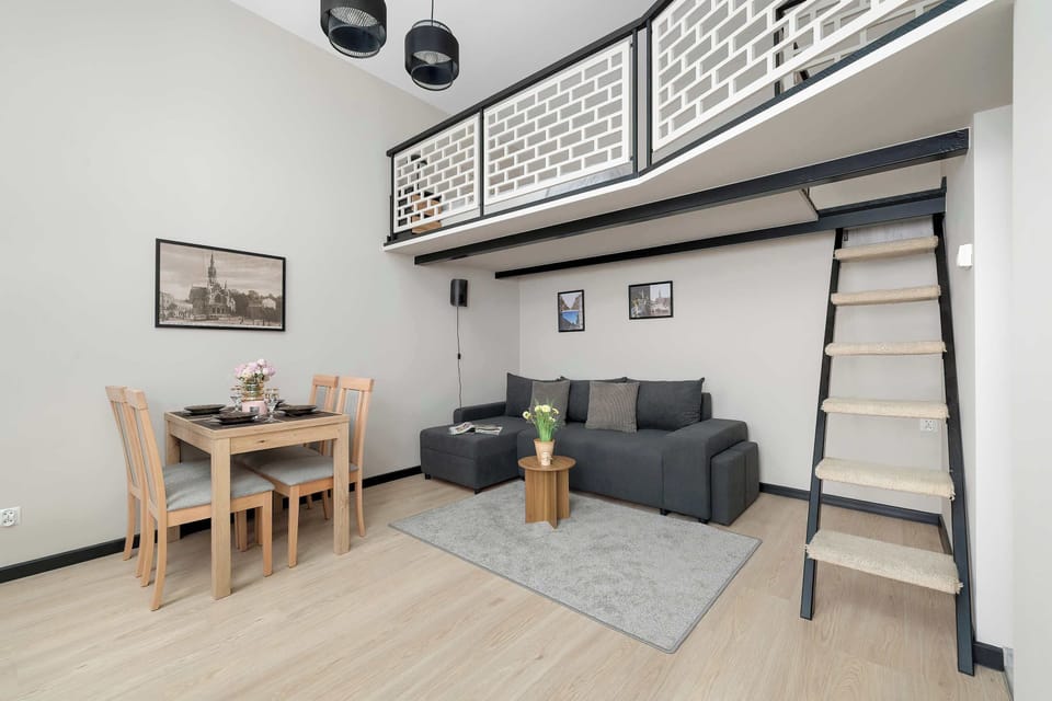 The open-plan apartment layout showcases a living area with a dark sofa and a wooden dining set. A black metal ladder leads up to an elevated mezzanine sleeping area.