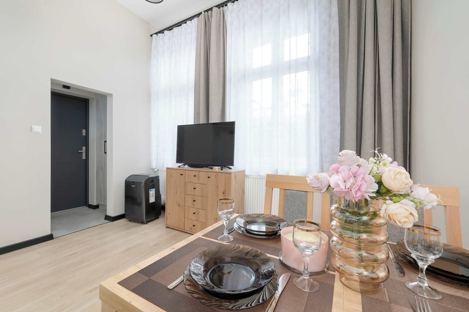 A dining table is neatly set with plates, glassware, and a vase filled with pink flowers. The entrance door to the apartment and a TV unit are visible in the background.