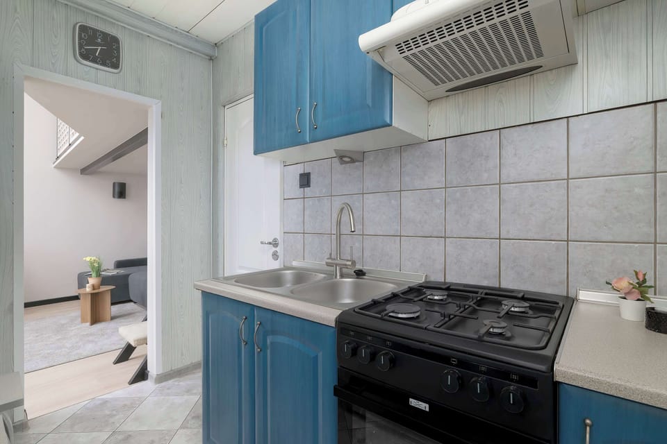 The kitchen area features classic blue cabinetry contrasted by tiled walls and a black gas stove with an oven. An open doorway leads the eye toward the brighter main living area.