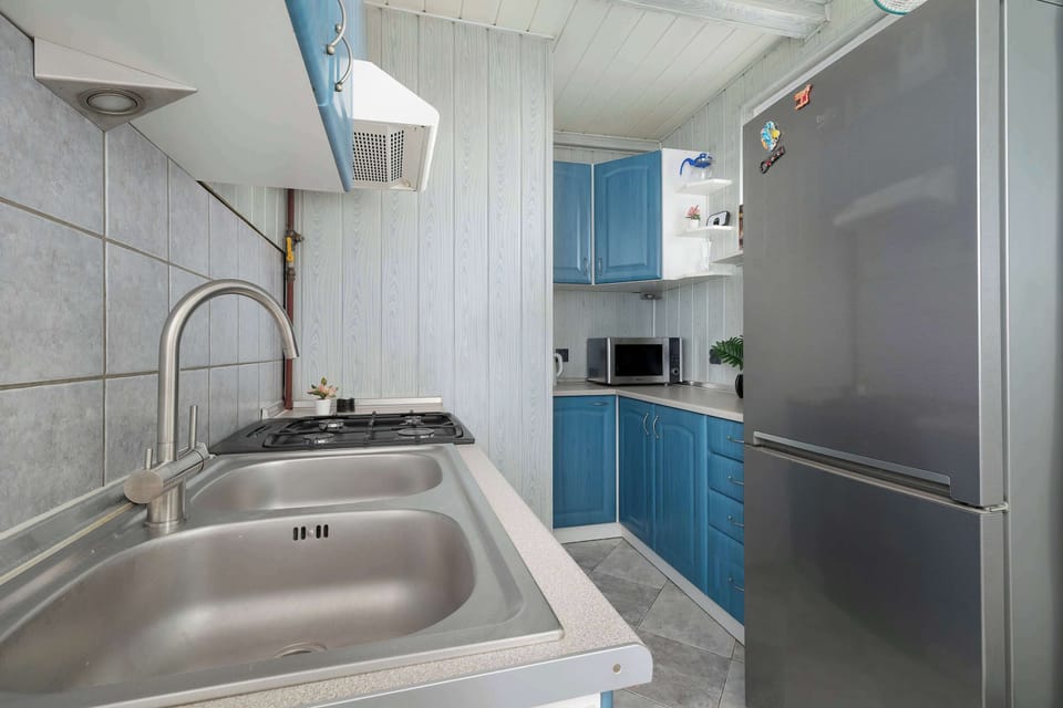 A compact kitchen corridor shows a stainless steel sink set into a light countertop. A large silver refrigerator stands opposite the blue cabinets, maximizing the narrow space.