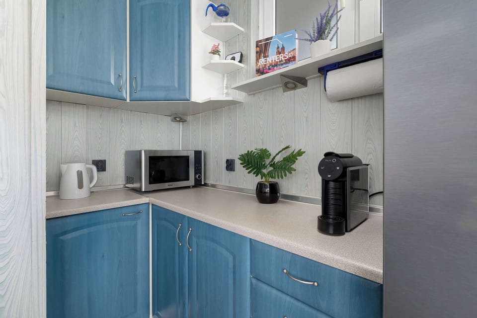 Bright blue kitchen cabinetry lines the corner of a functional cooking space. A microwave, coffee machine, and electric kettle sit on the light-colored countertop.