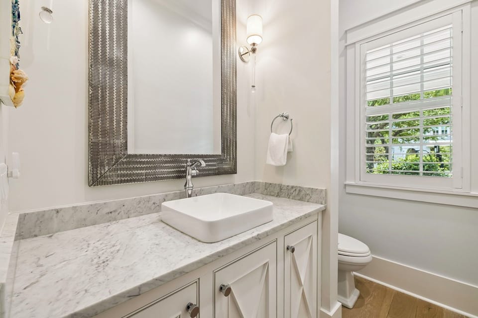 Step into this bright, elegant bathroom where marble countertops and modern fixtures create your perfect morning routine. Natural light pours in, making every detail—from the framed mirror to the plush towels—feel thoughtfully designed for your comfort.