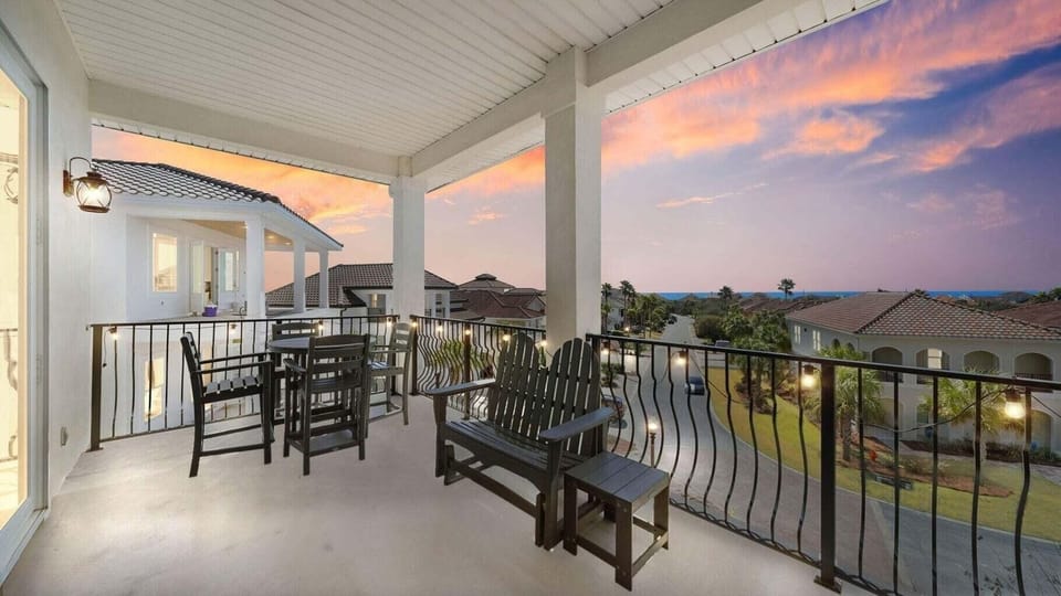 A covered balcony with dining and lounge seating overlooks a residential community beneath a vibrant sunset sky. The space provides a peaceful vantage point for evening gatherings.