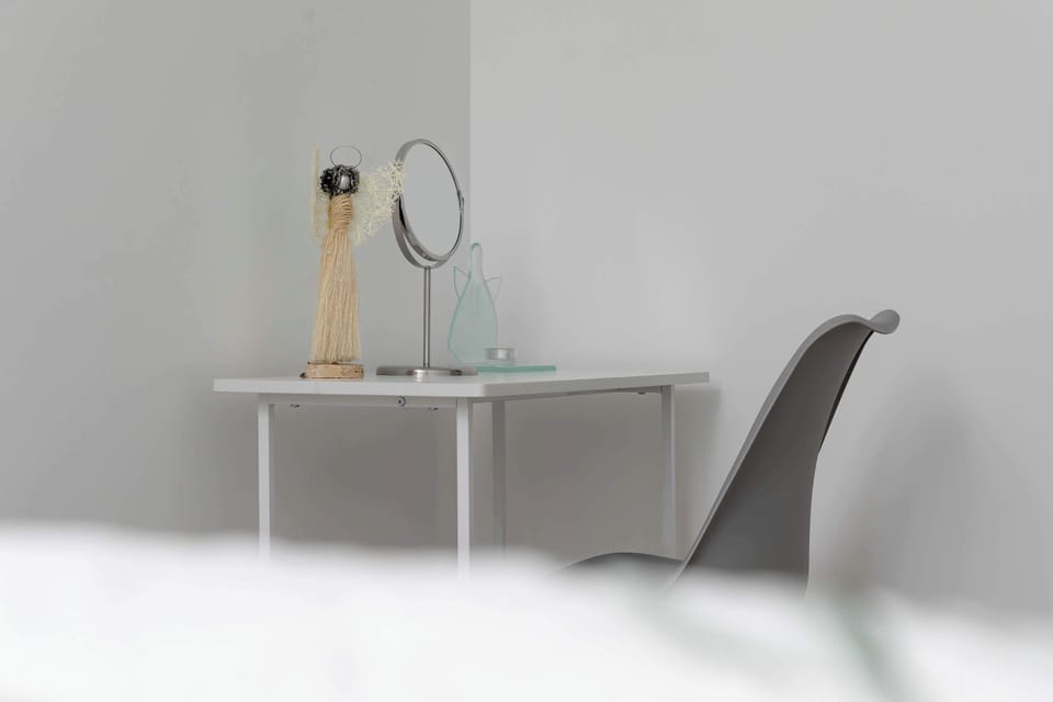 A close-up displays a simple white workspace setup containing a desk, a mirror, and a sleek black chair. The minimal background highlights the clean lines of the furniture.
