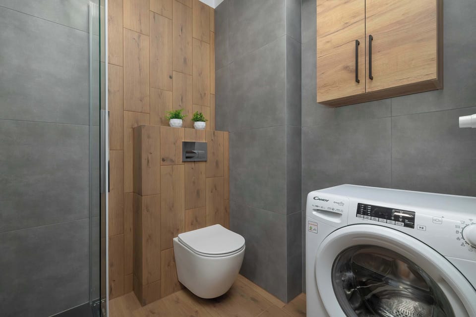 A utility corner in the bathroom features a front-loading washing machine tucked under a wooden counter next to the toilet. Light grey marble-patterned wall tiles cover the surrounding walls.
