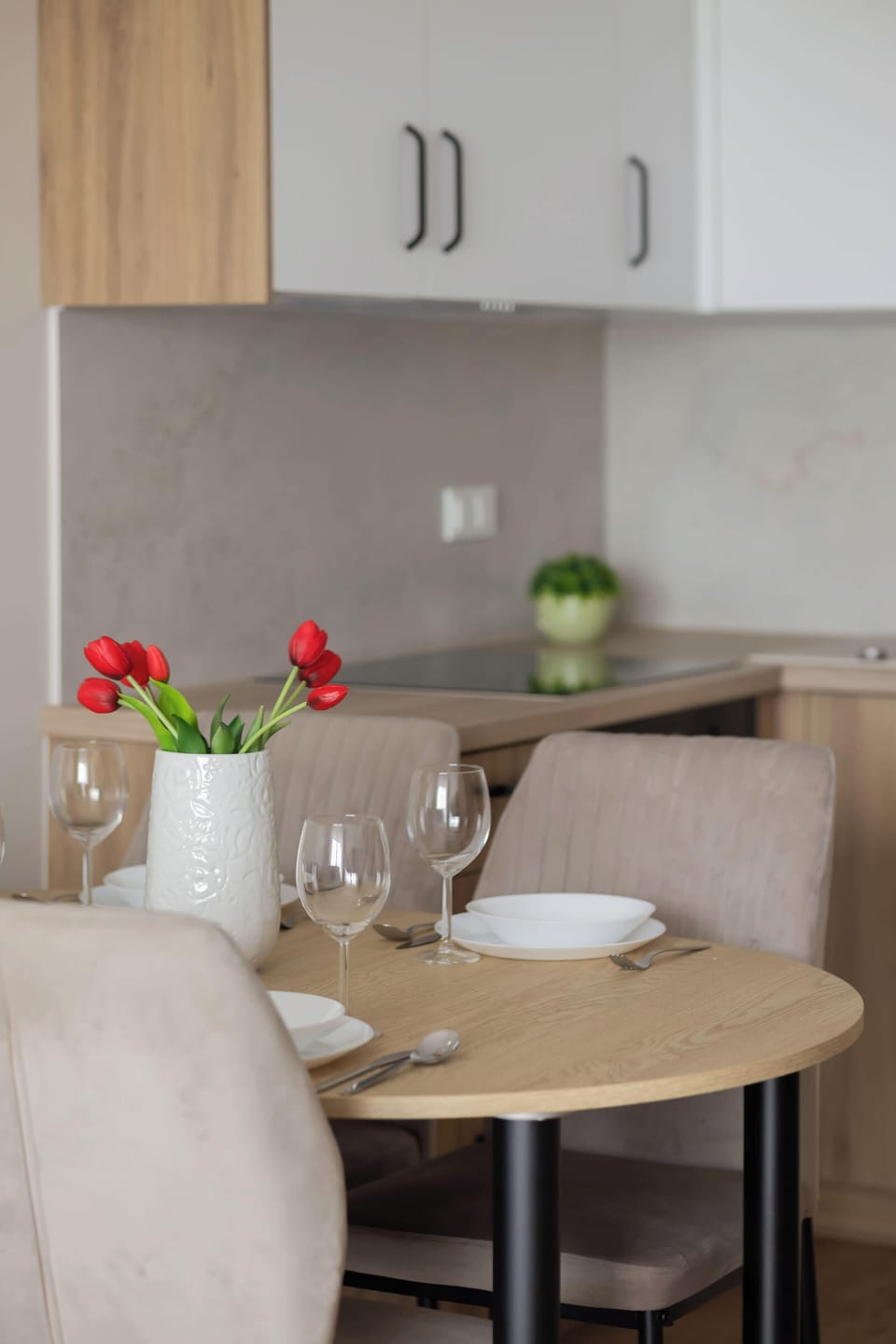 A close-up view focuses on a compact round dining table set with white plates. A small vase with red flowers sits on top beneath white cabinets.