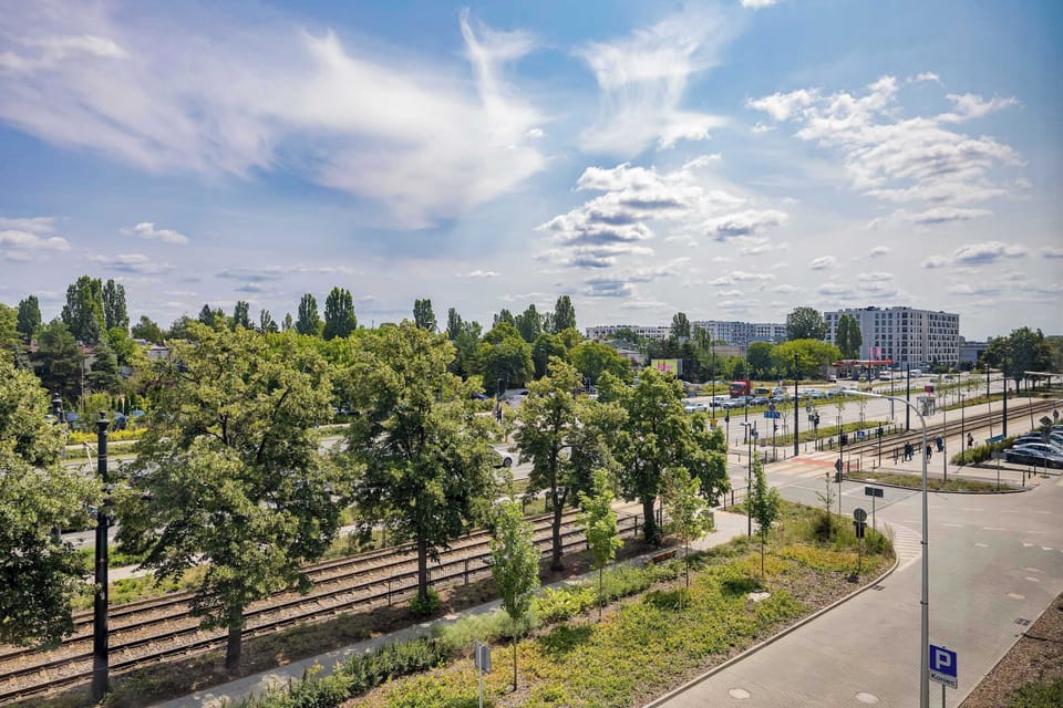 A high-angle view looks down from the balcony onto a landscaped city street lined with rows of green trees and tram tracks.