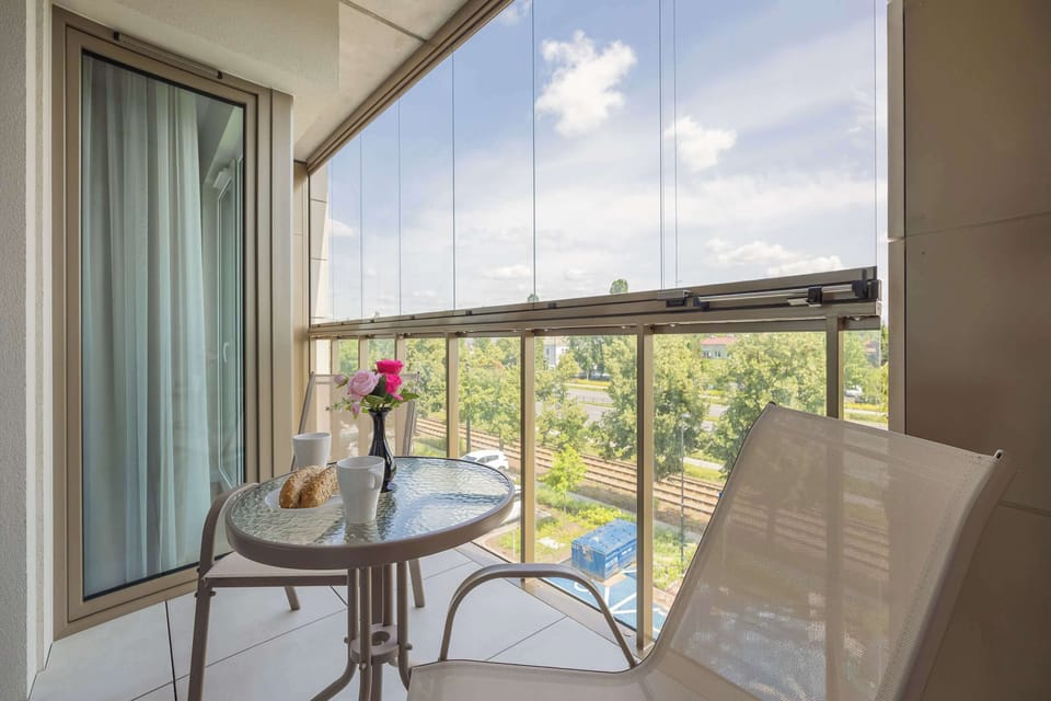 A glass-enclosed balcony is furnished with a round metal table and chairs. The elevated viewpoint looks down onto a green park filled with trees.