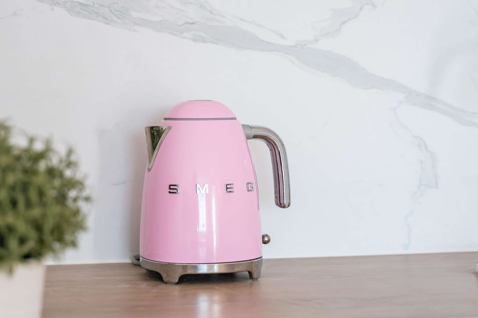 Le matin, la bouilloire rose SMEG chuchote déjà: sur le bois chaleureux et la crédence marbre, préparez un café cosy avant de partir — une touche rétro qui réveille la cuisine.