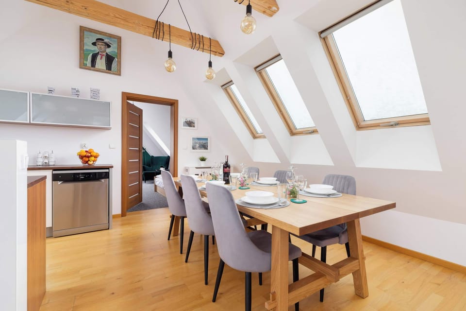 Bright open-plan dining area with a wooden dining table for four, large skylights, exposed wooden beams, and a modern kitchenette in the background.