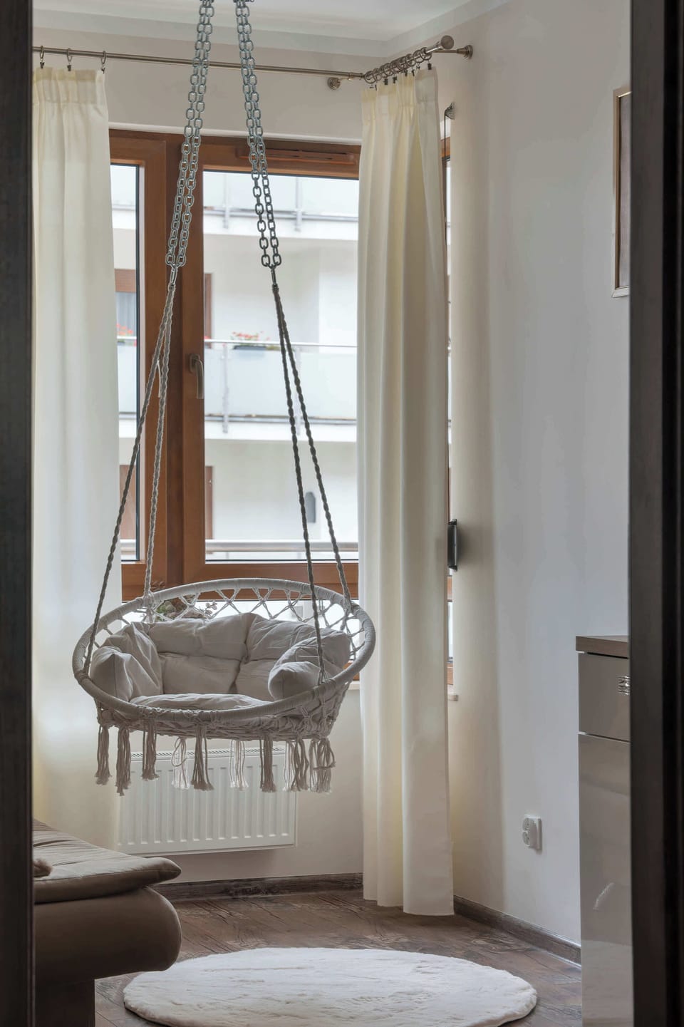 A focused vertical shot captures a distinctive hanging wicker swing chair suspended from the ceiling near a window. Sheer white curtains frame the window, letting in soft natural light.
