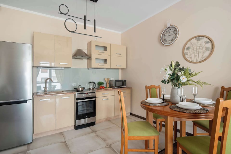 A functional kitchen area includes light wood cabinetry, a stainless steel refrigerator, and an oven with a stovetop. A round wooden dining table set with plates and a floral centerpiece stands adjacent to the cooking area.