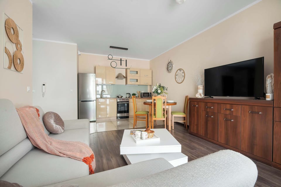 A wide-angle view of the living space captures the entire layout, featuring the corner couch, a white coffee table, and a dark wooden dresser under a large TV. The kitchen area is visible in the far background, seamlessly integrated into the room.