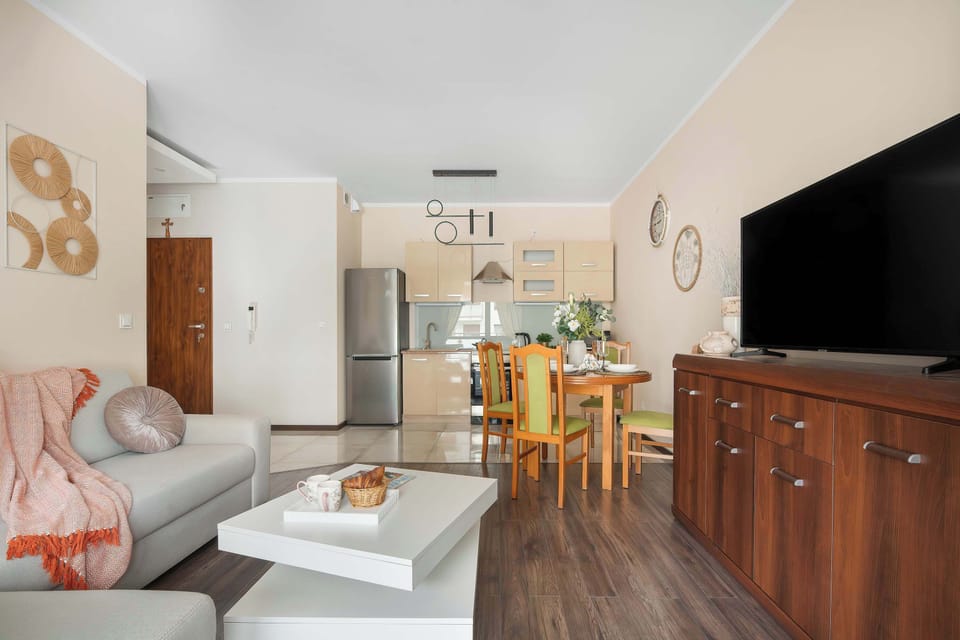 A perspective of the main living room shows the gray sofa with a decorative throw blanket facing toward the open kitchen area. A wooden dining table sits between the living space and the kitchen cabinets in the background.