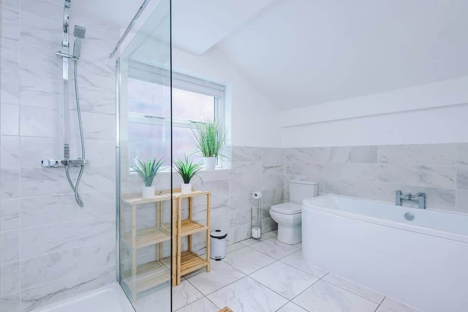 Bright, modern bathroom with marble walls, white bathtub, glass shower, and vanity unit. A wooden shelf with plants adds warmth, while natural light from the window creates a fresh, spa-like feel for guests to unwind in comfort.