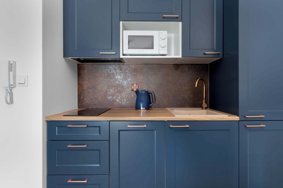 The compact kitchenette features built-in navy szafki, a wooden countertop, and an integrated white microwave. A black kettle rests on the counter.