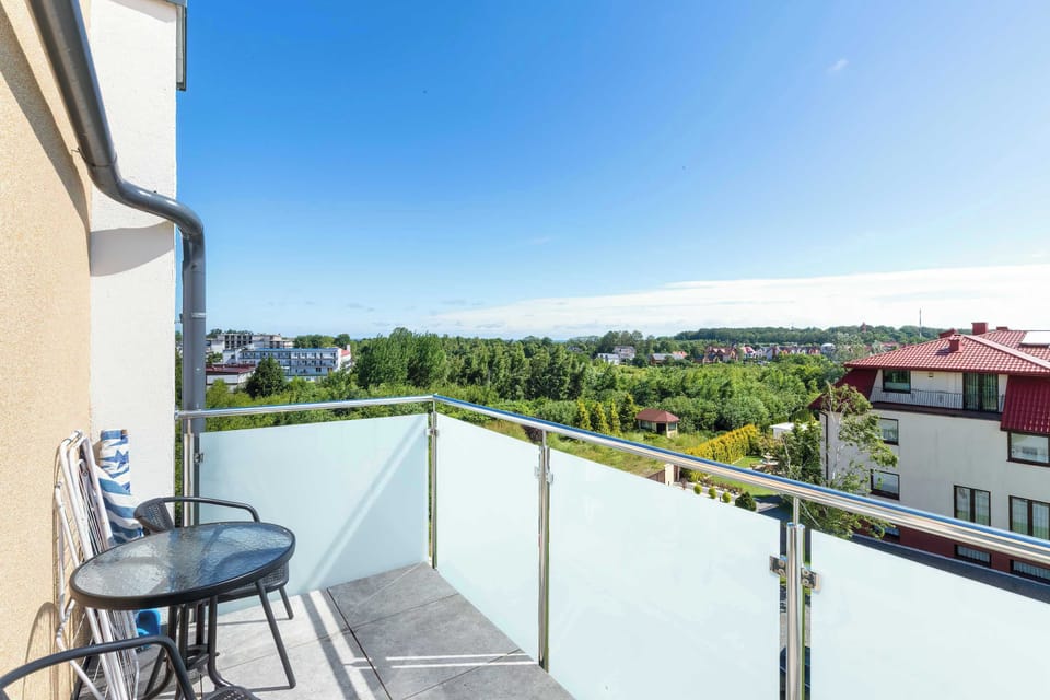 A glass-paneled balcony is furnished with a small patio table and two woven chairs. The elevated viewpoint looks over a lush green horizon.