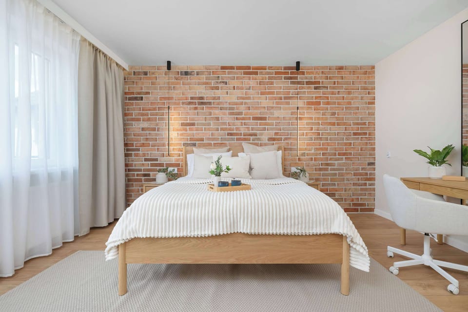 A serene bedroom layout displays a neatly made bed positioned directly in front of an exposed brick wall. A dedicated workspace with a white chair and desk sits quietly in the corner.