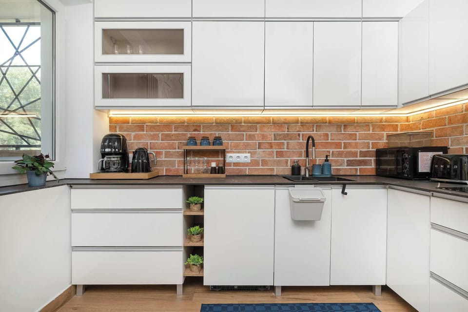 A bright kitchen workspace offers plenty of counter space under a large window. White minimalist cabinets are complemented by a warm brick wall and various small kitchen appliances.