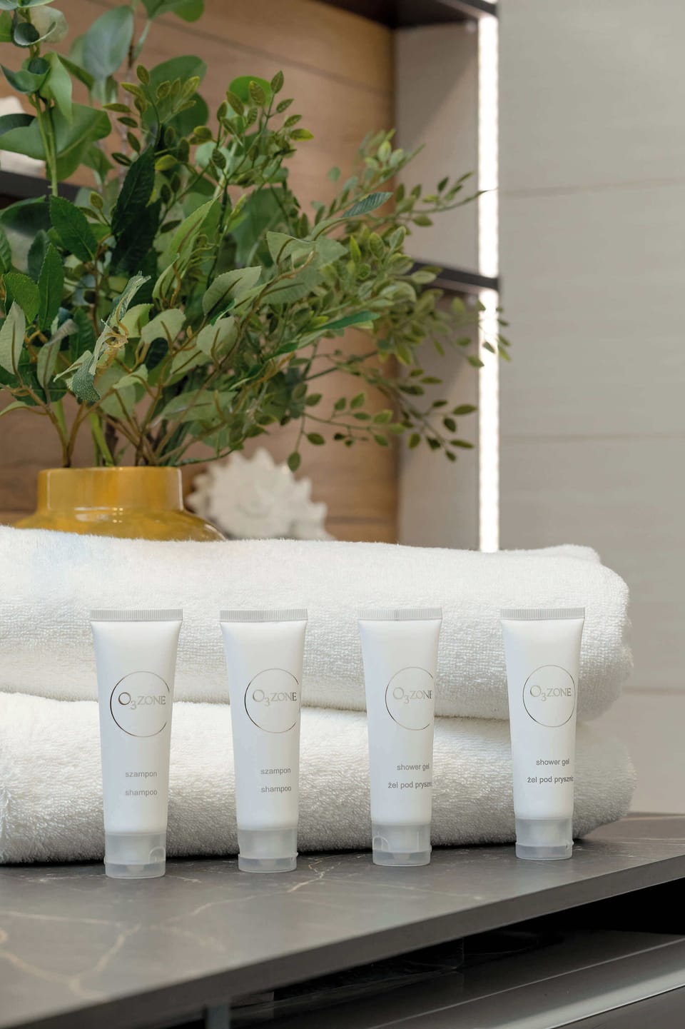 A small arrangement features rolled-up white towels and a set of miniature toiletry bottles placed neatly on a dark shelf. Green foliage in the background adds a refreshing touch of nature.