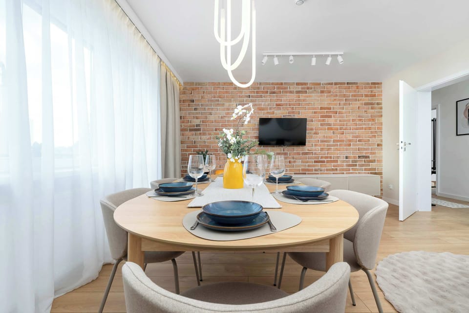 A circular dining table surrounded by upholstered chairs sits perfectly in front of a brick feature wall. A large window lets in ample daylight, making the dining space feel warm and inviting.